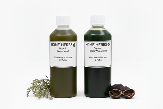 Two 500ml herbal tincture bottles labeled HOME HERBS Organic Black Walnut Hulls and Organic Wormwood, arranged on a white background with green/black walnut hulls and green wormwood sprigs.
